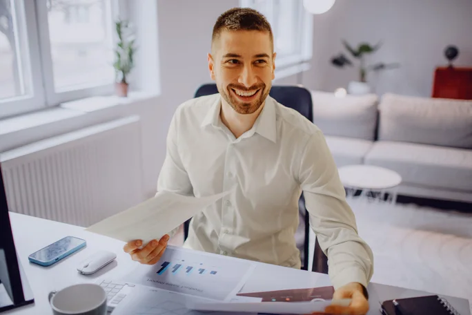 Eine Person im weißen Hemd sitzt an einem Schreibtisch, hält Papiere in der Hand und analysiert ausgedruckte Diagramme. Der Arbeitsplatz befindet sich in einem modernen, hellen Raum mit natürlichem Licht und minimalistischer Einrichtung. Auf dem Schreibtisch liegen ein Smartphone und weitere Büroartikel, was die professionelle Umgebung unterstreicht.