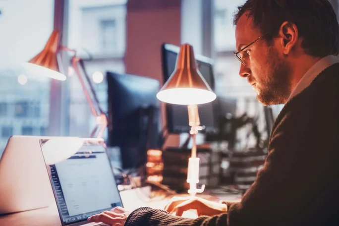 Ein Mann arbeitet nachts im Büro am Laptop unter Schreibtischlampen. Produktivität und Fokus auch zu später Stunde. Auri Häufig gestellte Fragen Seite Geschäftskunden.