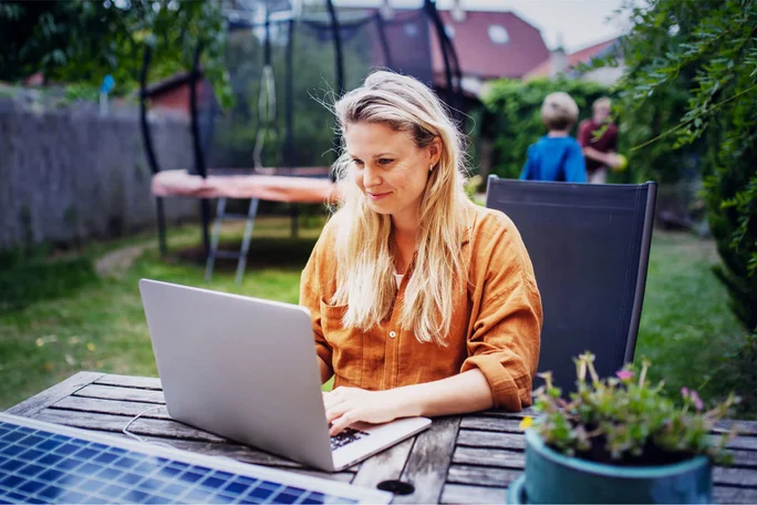 Eine Frau sitzt im Garten und arbeitet am Laptop, während ihre Kinder spielen. Nachhaltige Energie und Flexibilität im Alltag.