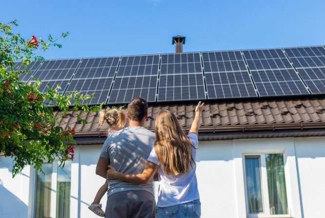 Eine junge Familie steht vor ihrem Haus und blickt zu den Solarmodulen auf dem Dach hinauf. Die Frau deutet auf die Module und unterstreicht so deren Bedeutung. Es ist ein sonniger Tag mit strahlend blauem Himmel, und das Haus wirkt modern und hell. Das Bild vermittelt ein Gefühl von Nachhaltigkeit, umweltbewusstem Leben und familiärem Zusammenhalt.